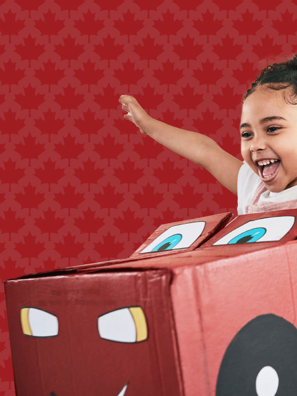 A child laughs and extends her arms while sitting in a cardboard car.